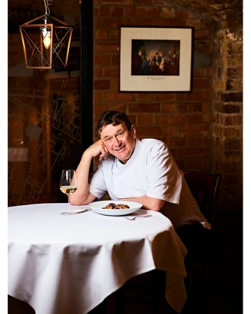 Portrait of a smiling male chef at a table with white tablecloth, wine, and plate of food inside a brick-walled restaurant.