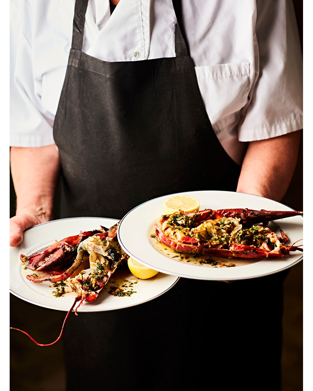 Chef holds two plates with cooked lobster halves topped with herbs and lemon slices, presented indoors under bright lighting.