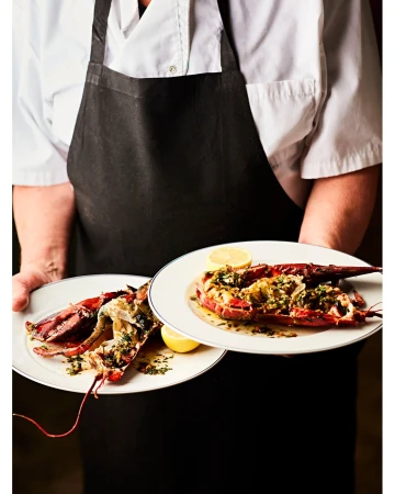 Chef holds two plates with cooked lobster halves topped with herbs and lemon slices, presented indoors under bright lighting.