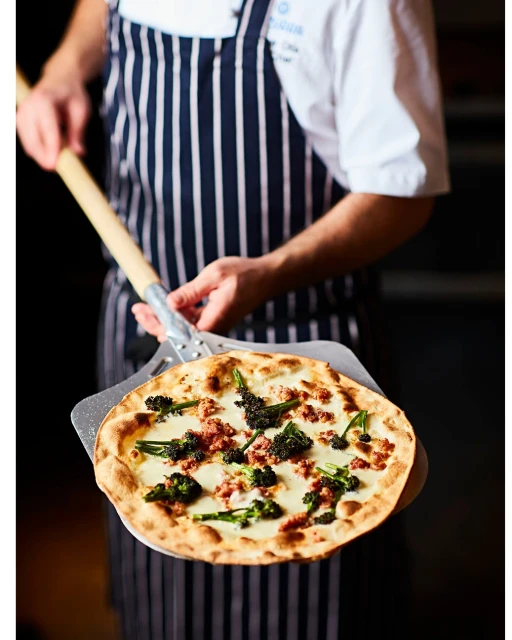 A chef in a striped apron holds a pizza peel with a freshly baked pizza topped with cheese, sausage, and greens inside a restaurant.