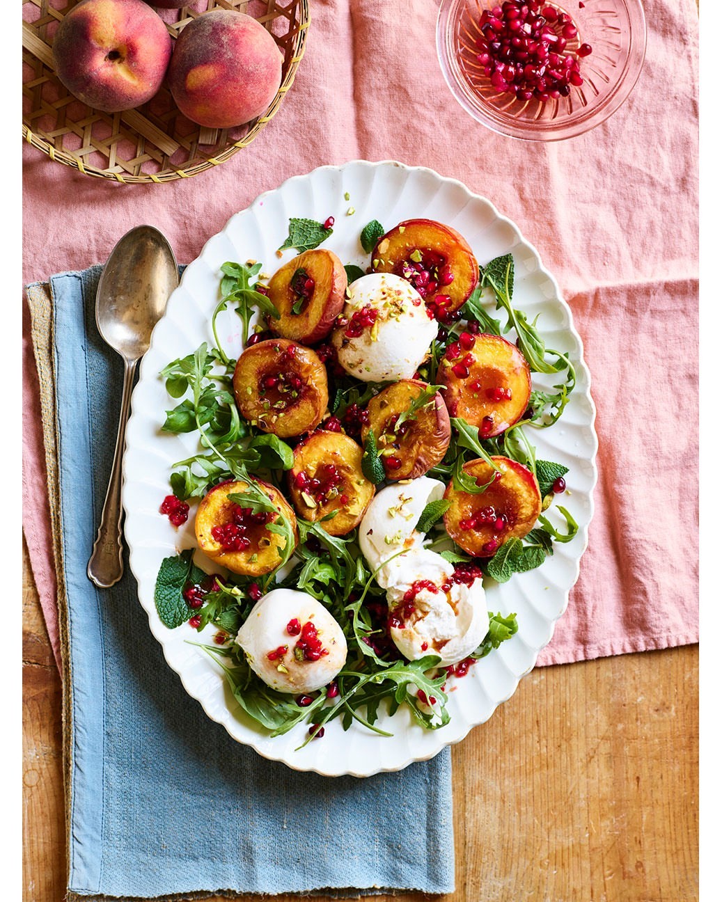 Overhead shot of a white platter filled with grilled peach halves, arugula, fresh mozzarella, and pomegranate seeds, set on a tablecloth.