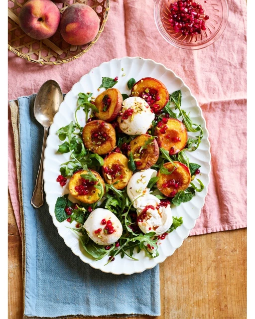 Overhead shot of a white platter filled with grilled peach halves, arugula, fresh mozzarella, and pomegranate seeds, set on a tablecloth.