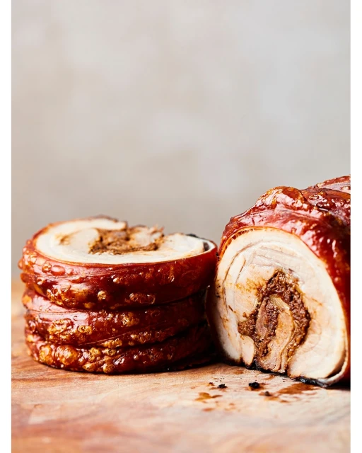 Close-up of a stack of sliced porchetta rolls on a wooden board, showcasing crispy skin and meat layers; neutral background.