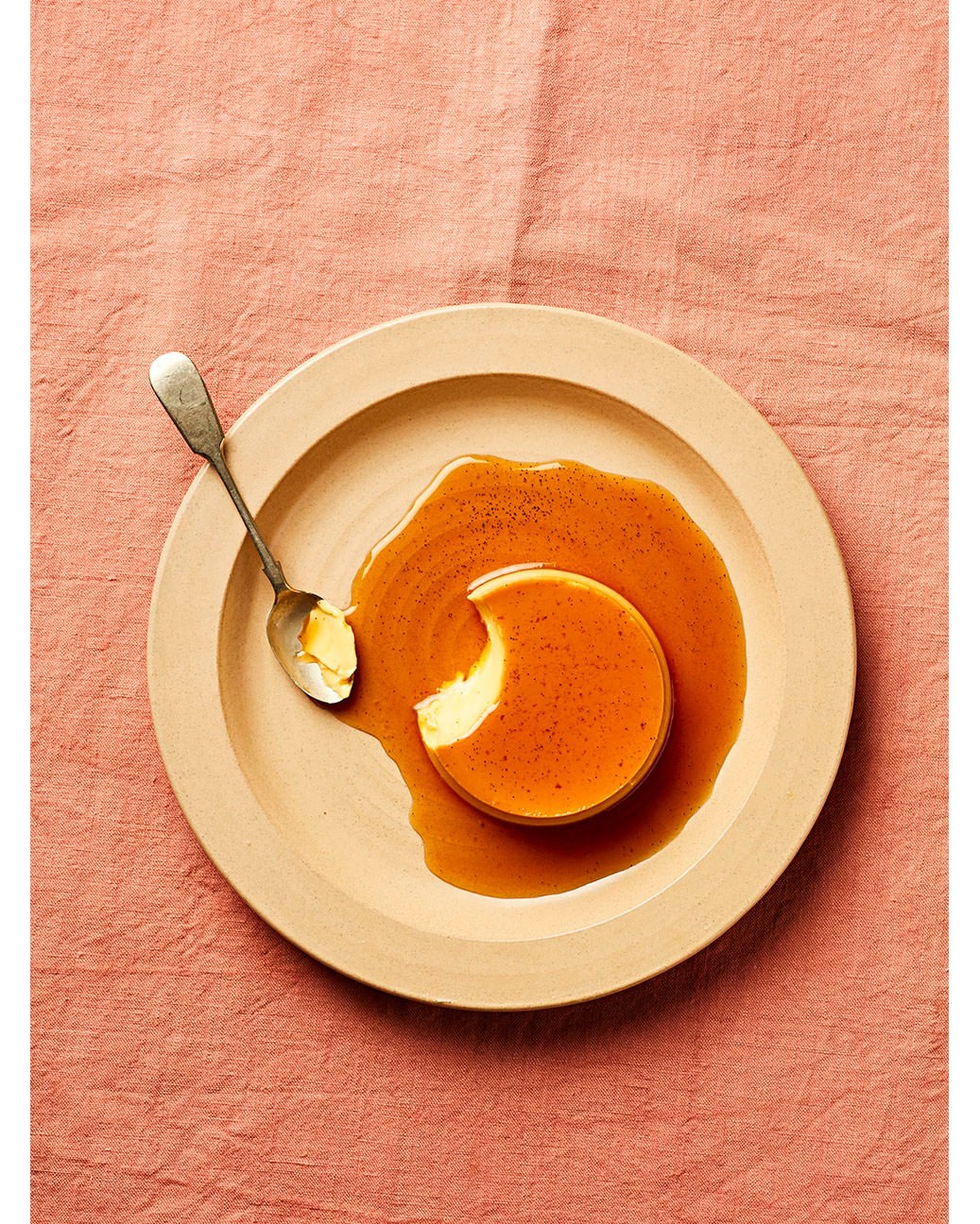 Overhead shot of a flan dessert on a plate with a spoon, featuring a smooth custard and caramel sauce on a textured pink surface.