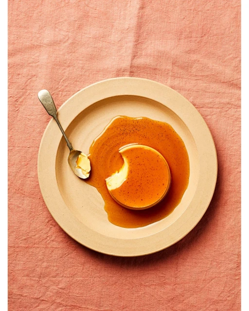 Overhead shot of a flan dessert on a plate with a spoon, featuring a smooth custard and caramel sauce on a textured pink surface.
