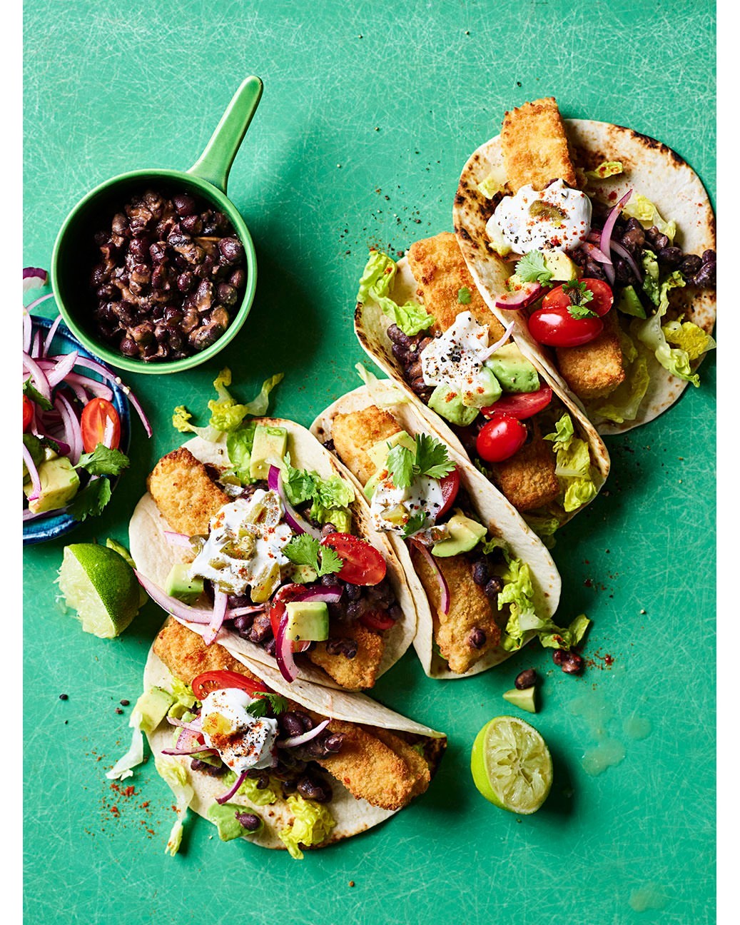Overhead shot of fish tacos with black beans, avocado, tomatoes, cilantro, and lime on a bright green surface. Food photography.