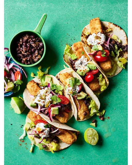 Overhead shot of fish tacos with black beans, avocado, tomatoes, cilantro, and lime on a bright green surface. Food photography.