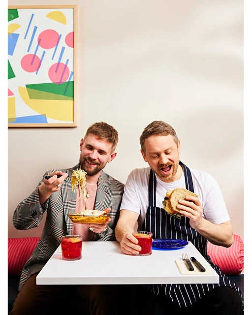 Two men at a white table enjoy food and drinks, one eating spaghetti, the other a sandwich, in a bright, indoor setting.