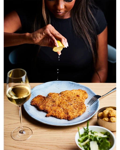 Woman squeezes lemon over breaded cutlet on a blue plate with wine, greens, and fried balls on a wood table in restaurant setting.