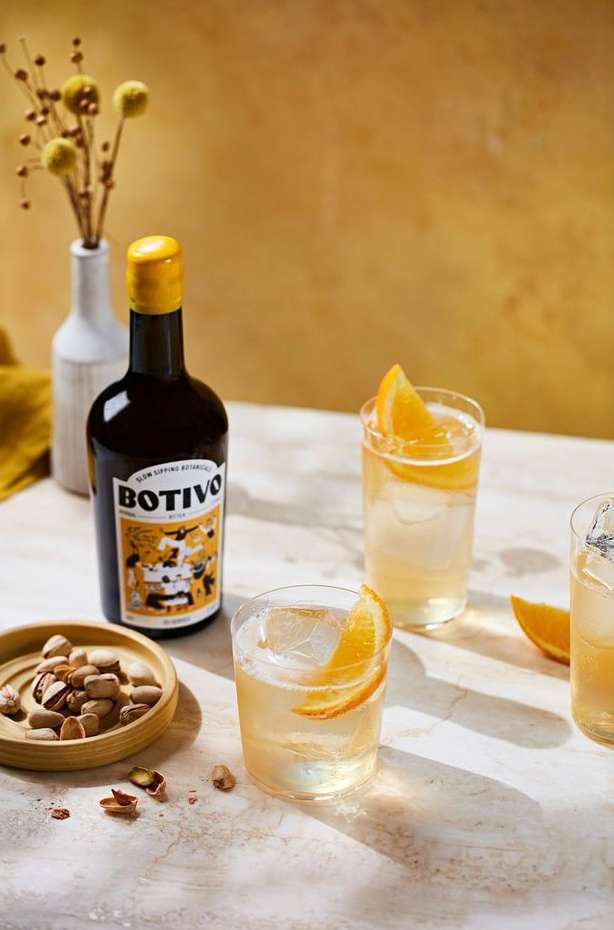 Elegant still life features BOTIVO aperitivo bottle, cocktail glasses with orange slices, and a small bowl of pistachios on a textured surface.