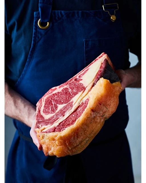 Chef in blue apron holds a large cut of raw red meat with visible fat marbling and bone in an indoor setting with neutral lighting.