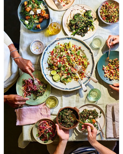 Overhead shot of a table set for a dinner party with multiple plates of colorful salads and vegetables being eaten by people.