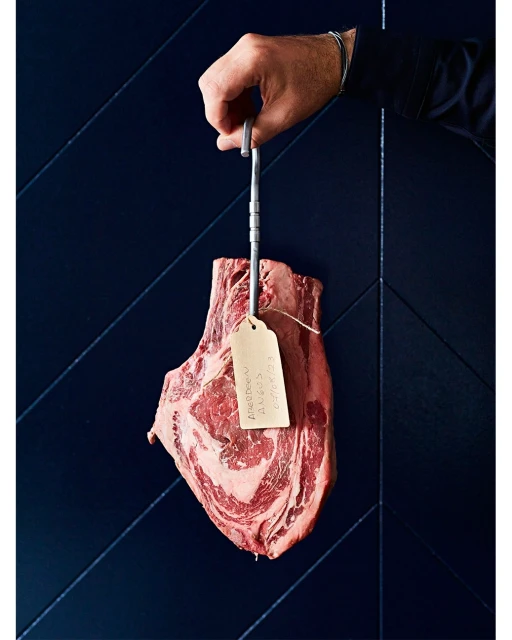 Bone-in ribeye steak hanging from metal hook against textured navy blue paneling, held by a hand wearing a dark long-sleeve shirt.