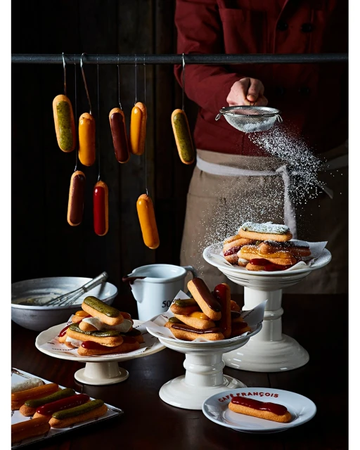 Overhead view of multiple colorful frosted eclairs displayed on cake stands and hanging from a metal rack in a studio setting.