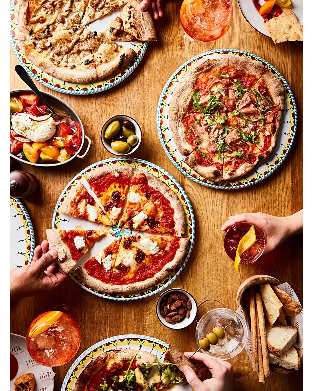Overhead shot showcases pizzas on patterned plates, accompanied by cocktails, olives, breadsticks, tomatoes, and hands reaching for food.