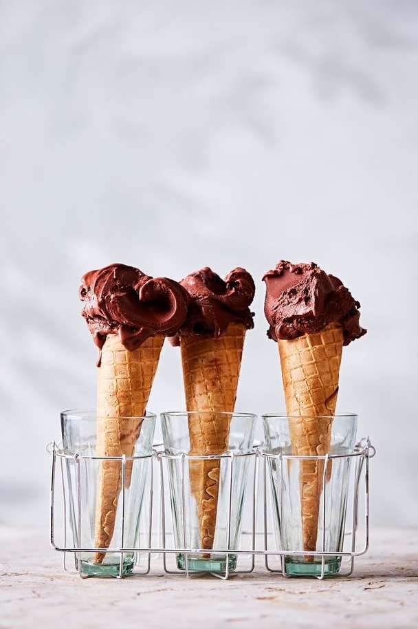 Three chocolate ice cream cones in waffle cones arranged in a wire rack with clear glasses against a neutral background.