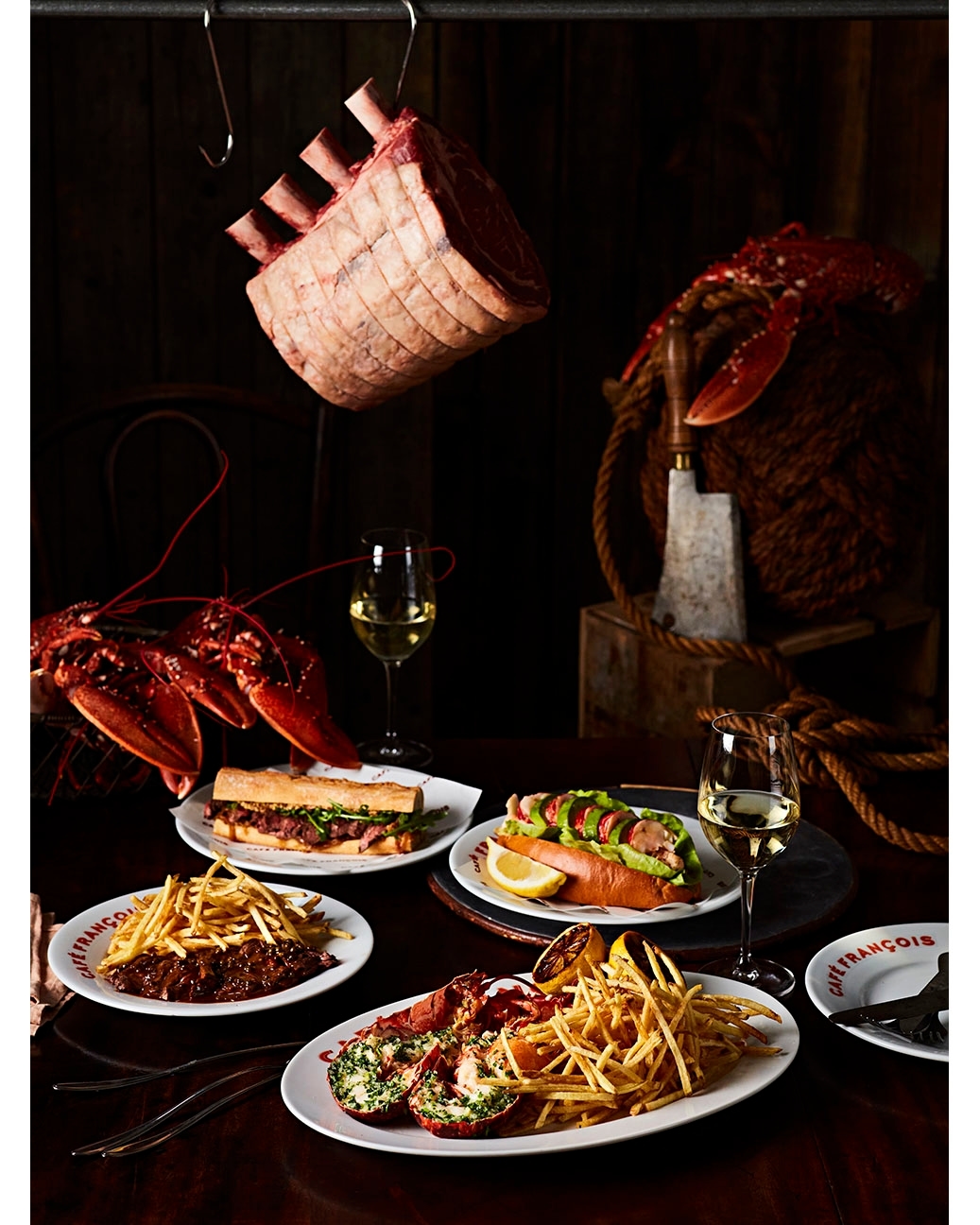 Overhead food photography on dark wooden table showcasing lobster, steak, hotdogs, fries, sandwiches, and white wine in wine glasses.