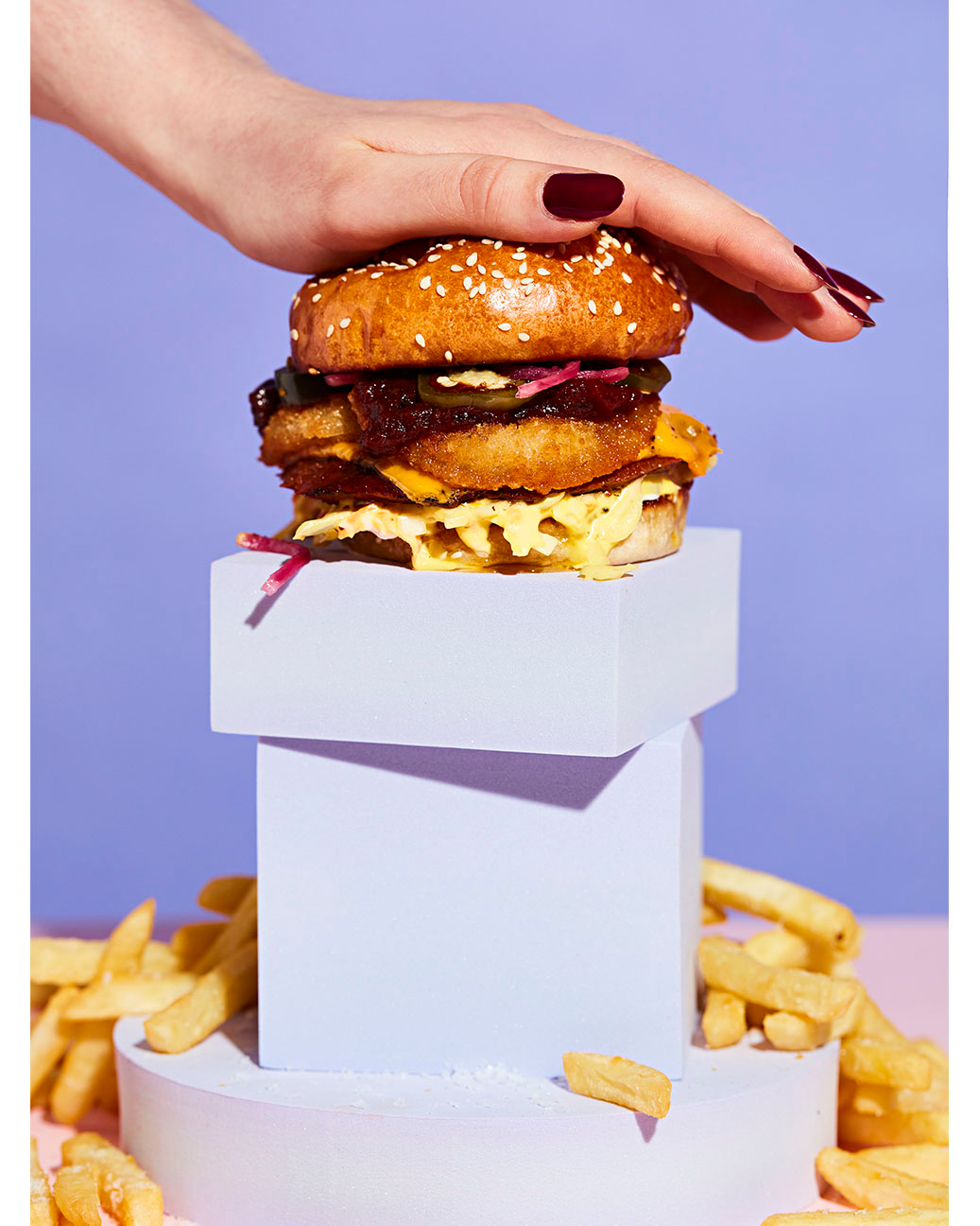 Close-up of a stacked burger topped with a hand, on a geometric platform with fries, against a gradient purple background.