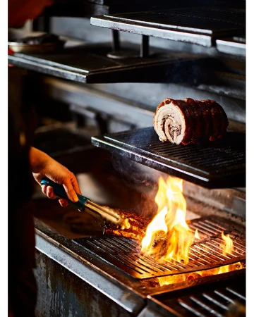 Chef grilling porchetta on a metal grate with flames in a dark restaurant kitchen, showcasing the culinary process.