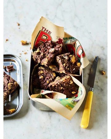 Chocolate fudge squares with honeycomb in a metal lunchbox lined with comic book paper, next to a dessert tray with a knife.