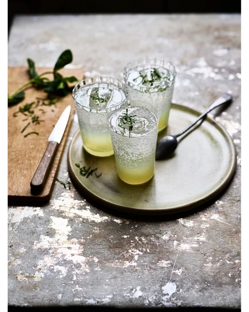 Three sparkling yellow drinks in clear glasses on a round green plate with a spoon and herbs on a wooden board, on a textured surface.
