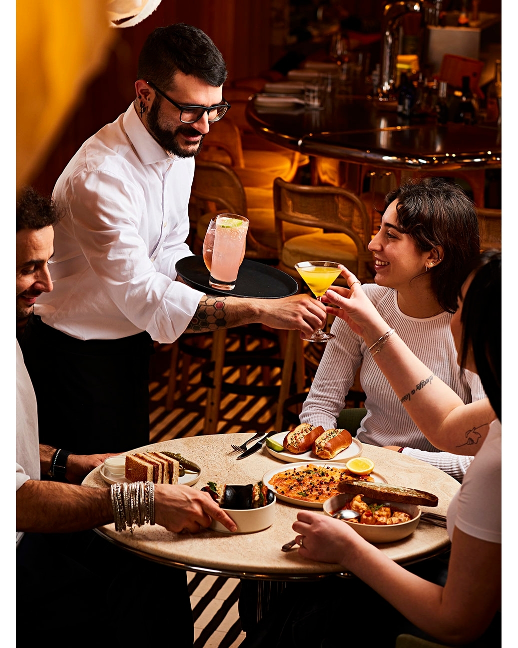 Waiter serving cocktails to a group of friends at a restaurant. The table is full of various food dishes and drinks.