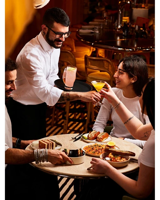 Waiter serving cocktails to a group of friends at a restaurant. The table is full of various food dishes and drinks.