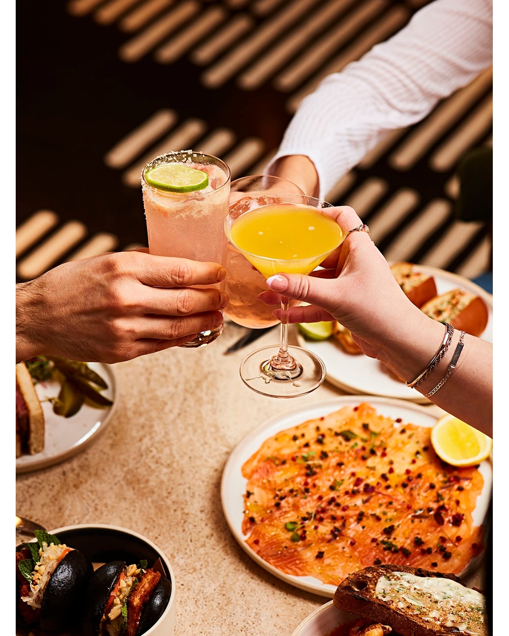 Three cocktails are being cheered in an indoor setting. There are various plates of food on the table, and there's a geometric background pattern.