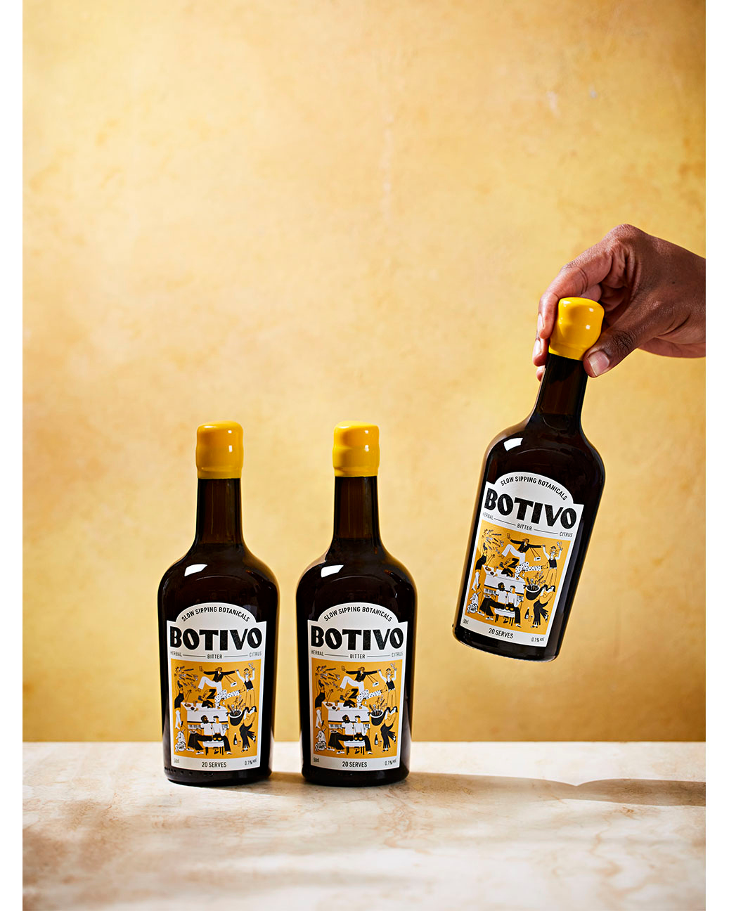 Three bottles of BOTIVO stand on a tan surface, with a hand holding one bottle against a mottled yellow background, advertising product.