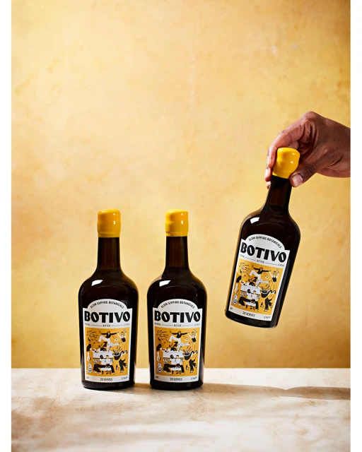Three bottles of BOTIVO stand on a tan surface, with a hand holding one bottle against a mottled yellow background, advertising product.