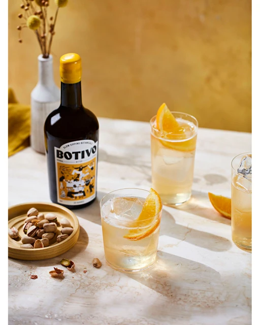 Refreshing cocktail glasses with orange slices, featuring a BOTIVO bottle and pistachios, set against a textured yellow backdrop.
