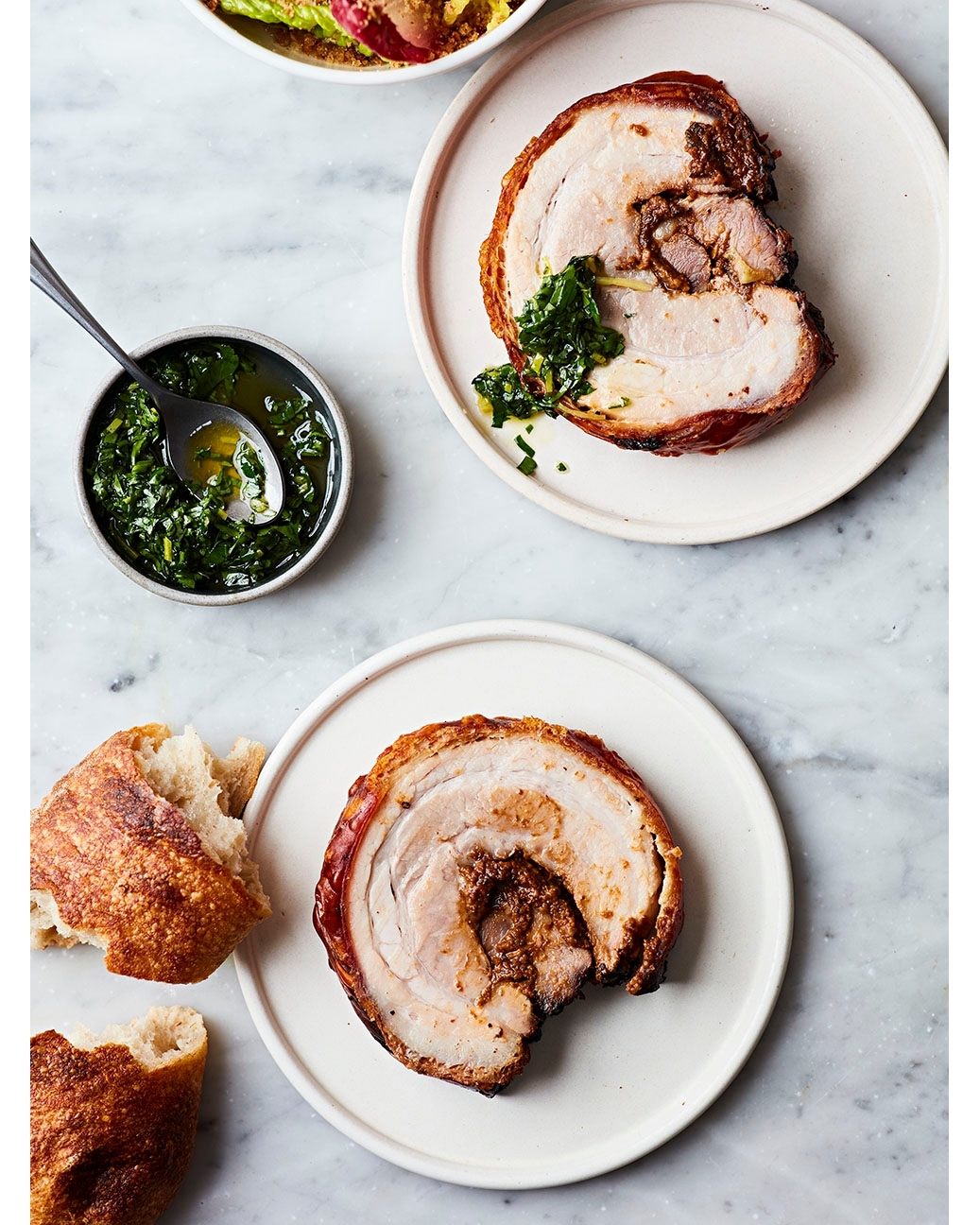 Overhead shot of two plates of porchetta with a side of bread and a small bowl of green sauce, all on a marble background.
