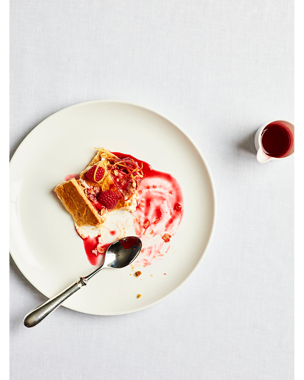 Overhead shot of a white plate with a dessert topped with raspberries and syrup, alongside a small silver spoon and creamer.