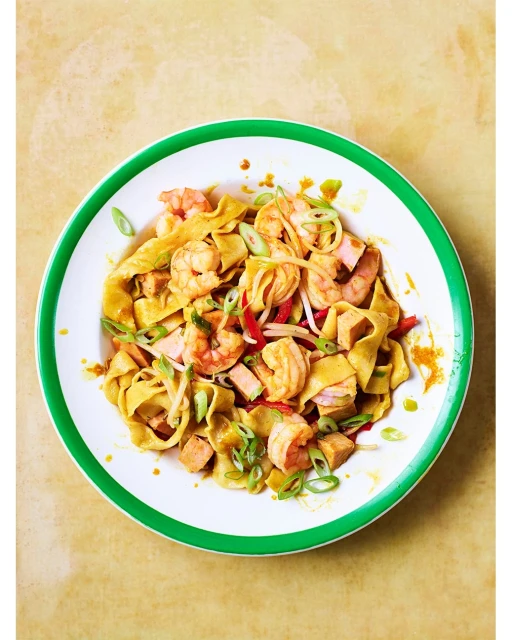 Overhead shot of shrimp and pasta dish with red peppers and green onions in white bowl with green trim on mottled gold surface.