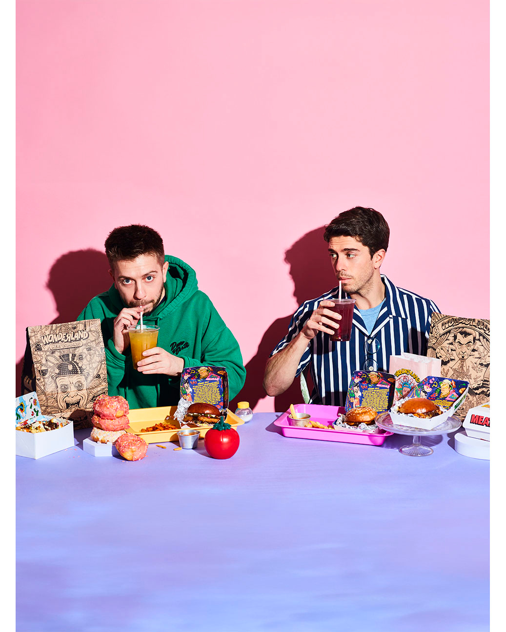 Two young men drinking from straws across a lavender table laden with burgers, donuts, and takeout containers against pink backdrop.