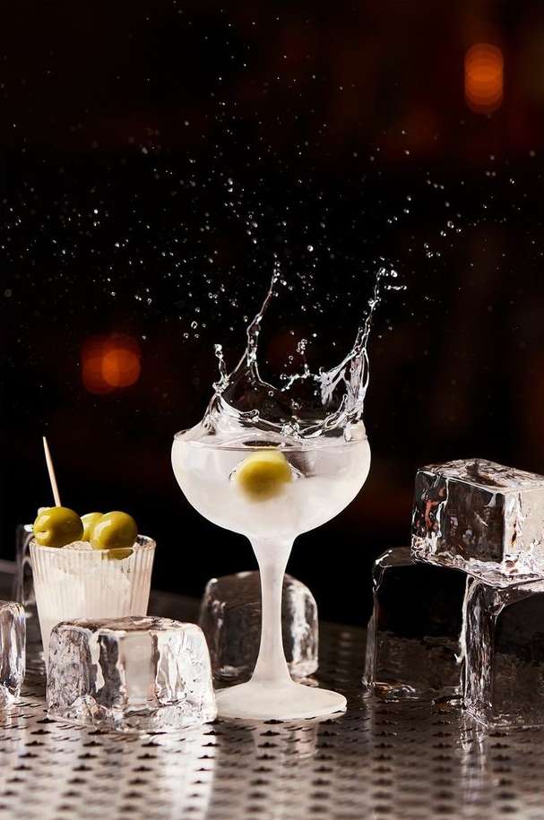Martini in coupe glass with olive garnish and water splash, cubes of ice and small glass of olives, on a metal bar top.