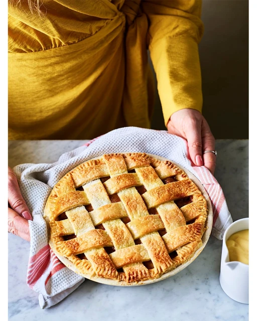 Overhead shot of golden-brown lattice-topped pie held by a person in yellow, with a white and red towel and custard on a marble surface.