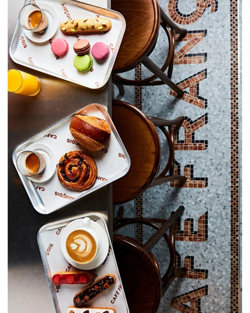 Overhead view of pastries, coffee, and orange juice served on metal trays at a cafe, with chairs and mosaic tile flooring.