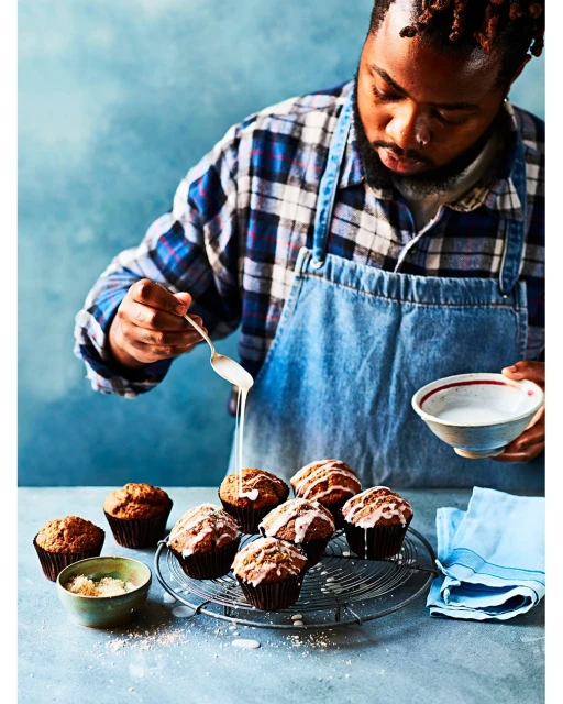 A man in a blue denim apron drizzles icing over several glazed muffins arranged on a wire rack in an indoor food photography setting.