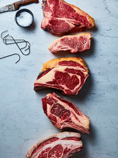Overhead shot showcasing a selection of raw beef cuts arranged on a light blue marble surface, accompanied by a bowl of salt and metal hooks.