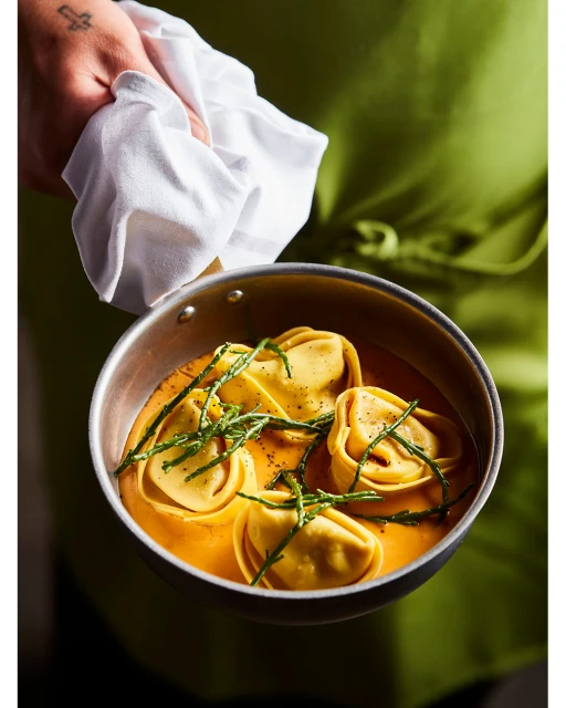 Close-up shot of a metal pan filled with tortelloni in orange sauce, garnished with green herbs, being held by a person in a green apron.