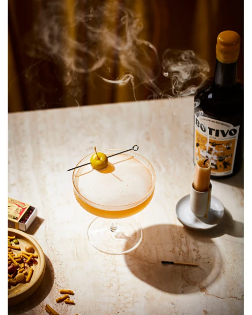 Cocktail in coupe glass with garnish, presented with smoke and vintage bottle on marble table, evokes a sophisticated, vintage mood.