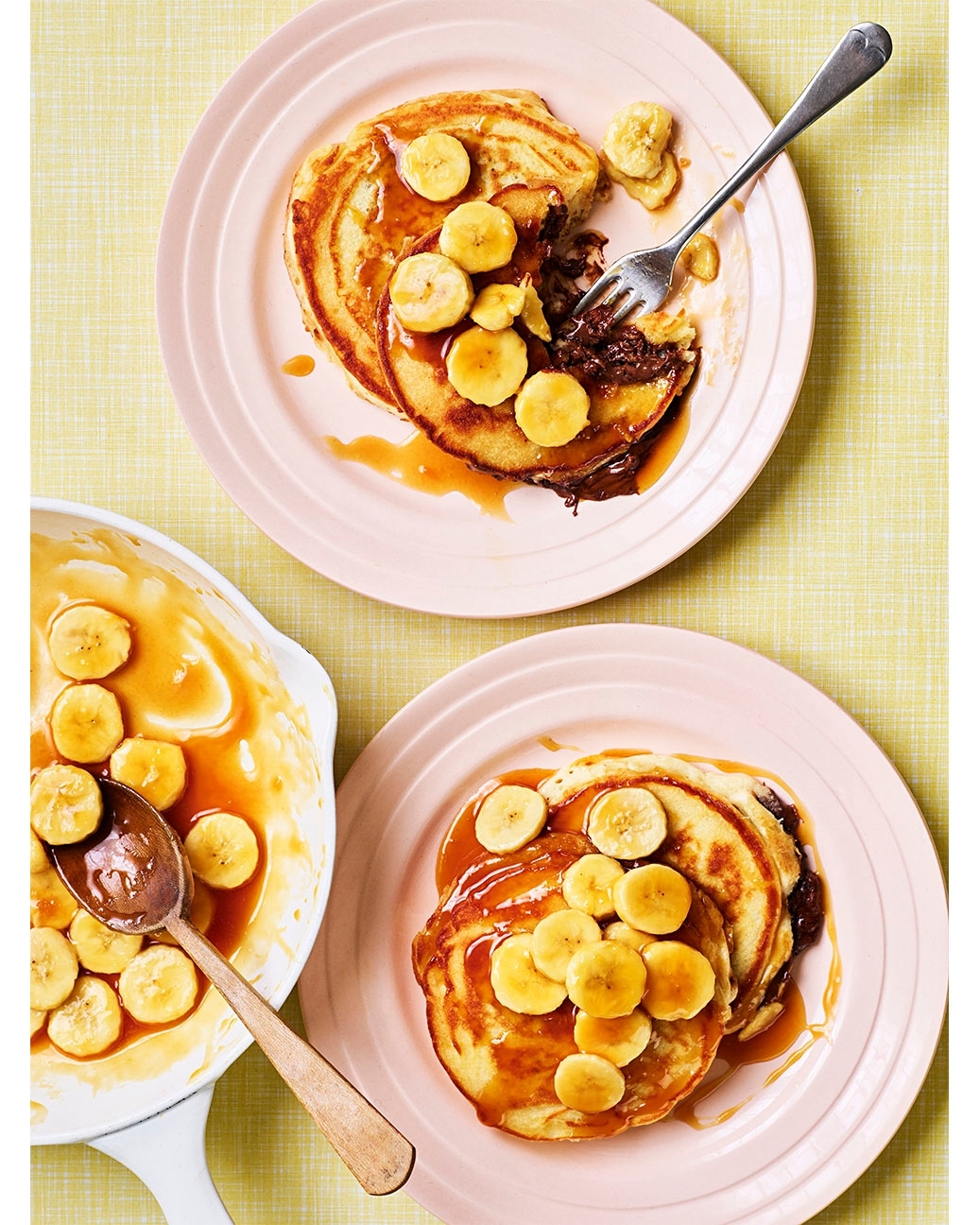 Overhead shot of two stacked pancakes with sliced bananas, drizzled caramel, and chocolate sauce on pink plates, with a fork and saucepan.
