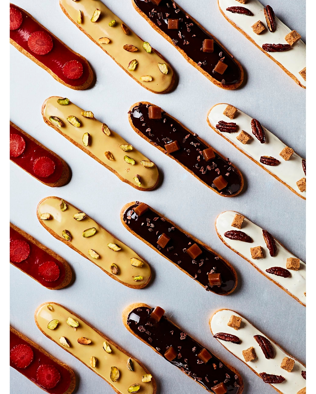 Overhead shot of an array of colourful eclairs. Flavours include strawberry, caramel, pistachio, chocolate and pecan in a clean food layout.