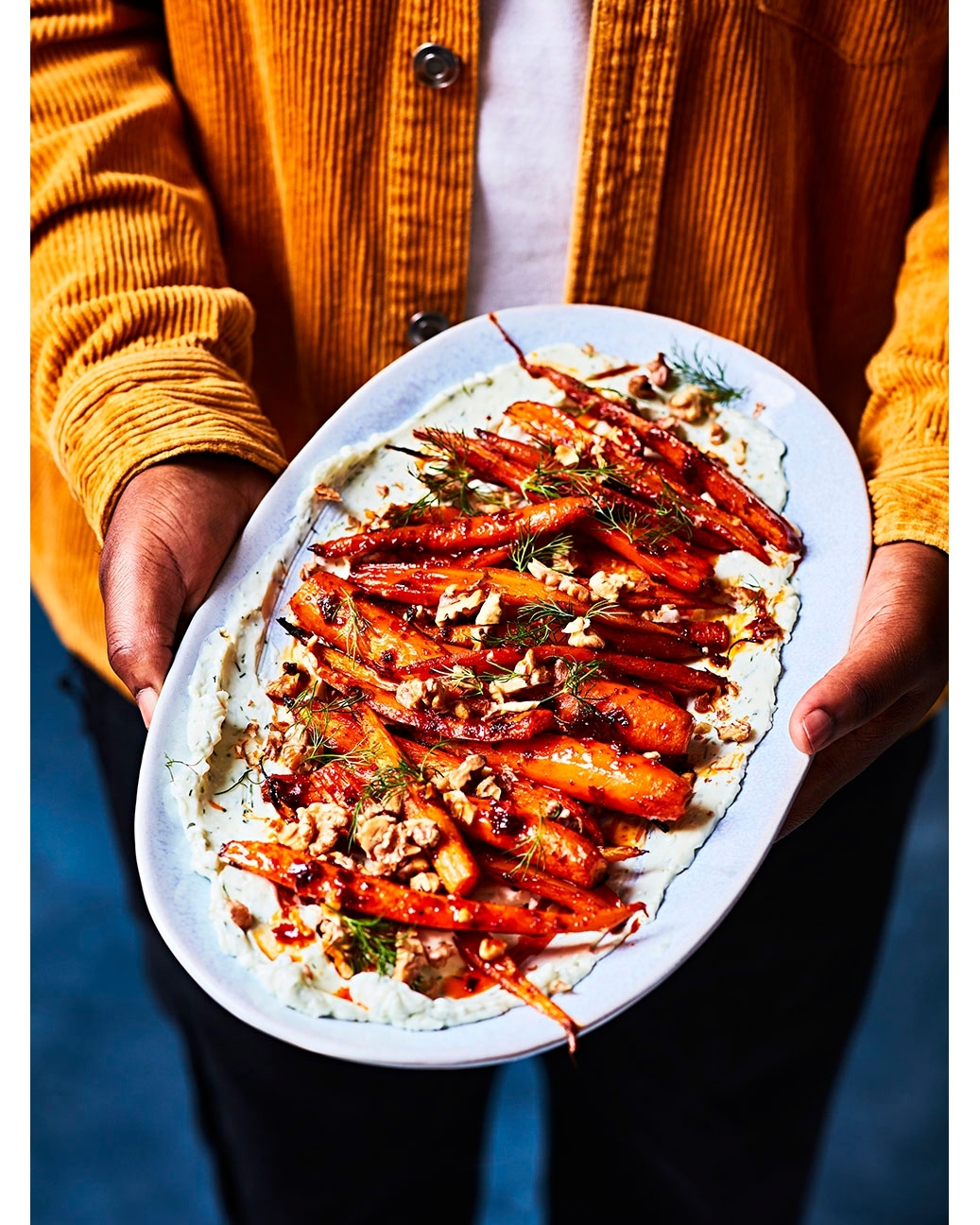 Person holding a platter of roasted carrots on white sauce with chopped nuts and dill, wearing a yellow corduroy jacket.