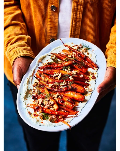 Person holding a platter of roasted carrots on white sauce with chopped nuts and dill, wearing a yellow corduroy jacket.