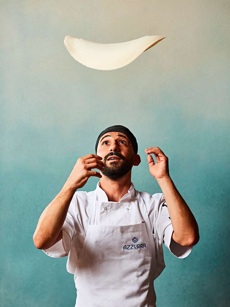 Chef in white coat tossing pizza dough in the air, against a mottled blue and beige backdrop. Food and beverage concept.