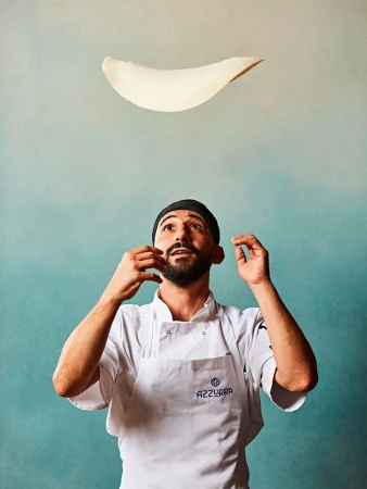 Chef in white coat tossing pizza dough in the air, against a mottled blue and beige backdrop. Food and beverage concept.