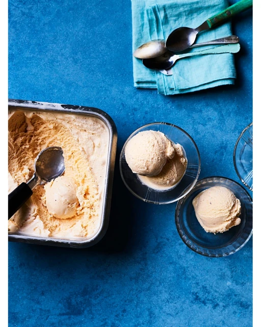 Overhead shot: metal pan of vanilla ice cream with scoop, two glass bowls with scoops, turquoise napkin with spoons on blue backdrop.