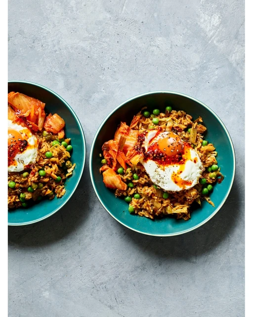 Two bowls of kimchi fried rice topped with fried eggs and garnished with peas sit on a grey textured surface, overhead shot.